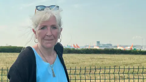 Mary Collett, a woman with white hair and sunglasses on the top of her head, is standing in front of a green field with Bristol Airport on the horizon.