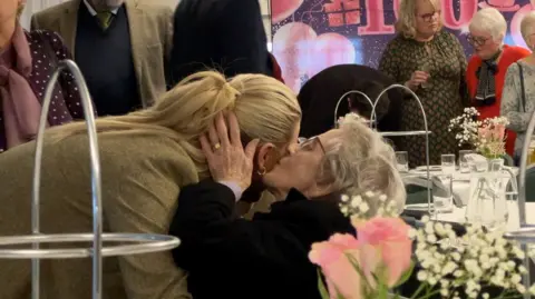 A woman leans down to be given a kiss on the cheek by Dorothy, who is embracing the side of the woman's head. Flowers can be seen on the tables nearby. 