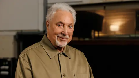 Sir Tom Jones, wearing a brown shirt, smiling. He has white hair and a white beard