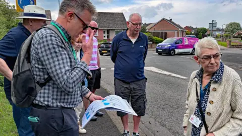 Ian Duncan Six people are standing on different spots on a pavement. One is looking down at a map the others are looking to the right. Behind them is a roundabout and some bungalows 