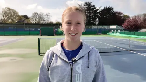 Daniella Britton wearing a grey and black hoodie and a blue t-shirt. She is stood on a blue and green tennis court and is staring at the camera smiling.