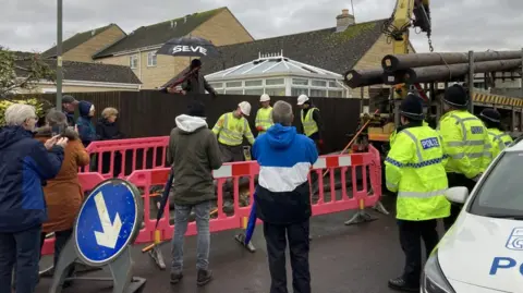 People, including residents and police officers, stand watching workmen preparing to install telecommunications poles.