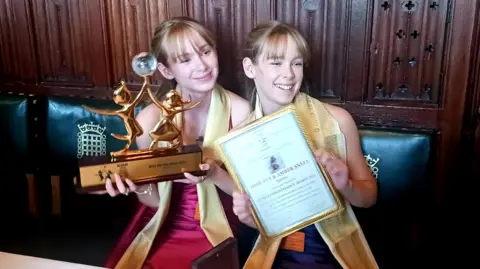 Zoe Snell A pair of teenage girls who are identical set of twins, both blonde and wearing gold scarves over their necks, holding up a gold award and a framed certificate. They are sitting on green-cushioned chairs with the Houses of Parliament logo.
