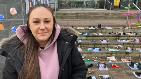 Stephanie Morrison, mother of a special needs child who attends Steps Ahead Nursery. She is wearing a pink hoodie underneath a black raincoat with a fur lined hood. She has long, straight brown hair and is looking at the camera. Behind her are the steps leading up to County Hall, where a demonstration has been set up with children's shoes lined up and handprints on the wall. 