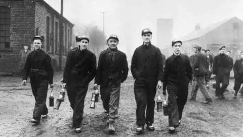 Getty Images Five miners, all carrying lamps walk towards the camera in dark overalls. They are all wearing hats. They are pictured in 1944.