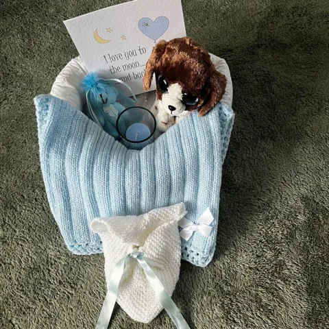 Humberside Police A small baby‑loss remembrance basket sits on a green textured carpet floor. There is a light blue knitted blanket wrapped around items including a soft toy dog, a small blue teddy ornament, and a tealight candle. A card with illustrations of a moon, stars, and hearts reads “I love you to the moon… and back.” In front of the basket lies a tiny white knitted pouch tied with a pale blue ribbon.