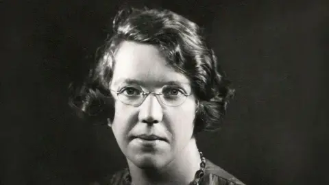This is a black and white picture. Miss Haining is wearing glasses and has short dark hair. She is wearing a necklace.