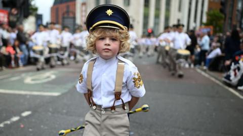 A young child with blonde curly hair marches in a band uniform 