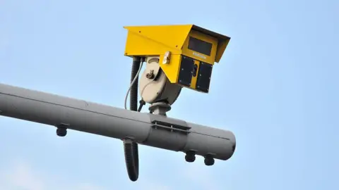 A close up of a yellow average speed camera resting on a grey pole, against a blue sky