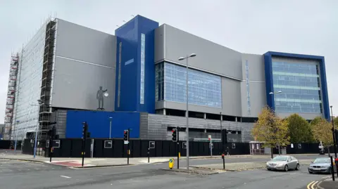 The former Ikea building in Coventry city centre is pictured. It is grey and blue and surrounded by scaffolding. It towers over nearby roads and cars.