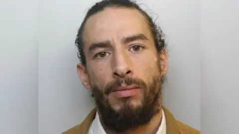 A custody picture of Cody. He has long dark hair pulled back from his face, dark eyebrows and a dark beard. He wears a white collared shirt and what appears to be a tan-coloured jacket. 