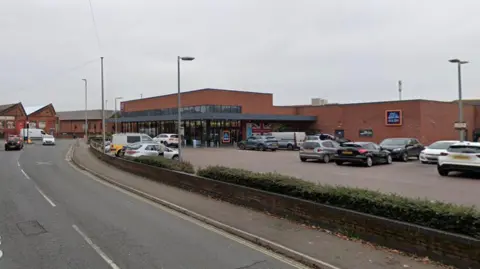 Google Maps street view of an Aldi store on Clifton Road in Gloucester. It is a red brick building with glass panels at the entrance. There is a car park to the side of the store and there are many cars parked up.