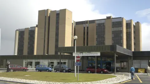 A hospital building in Inverness over a number of storeys with cars parked outside and people walking about nearby