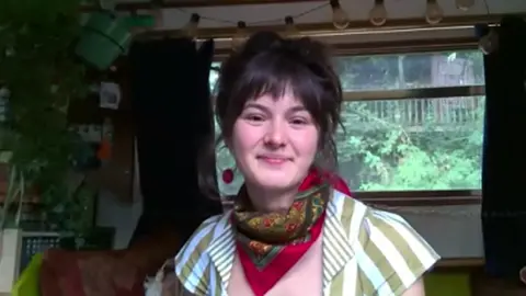 Anna, wearing a scarf and green and white striped top, sits on board a narrowboat with plants and artistic lights in the background.