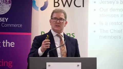 BBC Dr Andrew McLaughlin is wearing a dark blue suit with a multi-coloured tie. He is wearing glasses and is standing at a lectern in front of a power point presentation.
