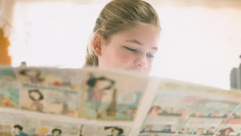 Getty Images A child holds open a comic and reads from it. The pages cover the lower half of her face as she gazes at them. Behind her is a large window, through which the sun is shining