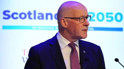 A bald man with glasses delivers a speech against white backdrop with Scotland 2050 on it