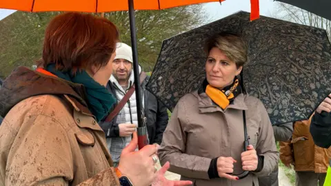 Two women are talking - the one on the left has her back to the camera - while the other - Emma Hardy, who has short brown hair, is looking at her. They are both holding umbrellas and wearing waterproof, brown coats.
