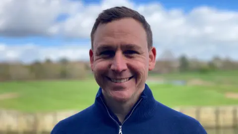Laura Coffey/BBC A man with short brown hair smiles at the camera. He is wearing a blue jumper. Behind him is the blurred background of a golf course with bunkers and a lake visible among the grass. 