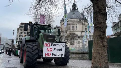 A green tractor is parked on a Westminster pavement under a grey winter sky next to a bare tree and in front of the QEII Centre. On the front of the tractor is a protest placard that reads No Farming, No Food, No Future. Pedestrians pass by.