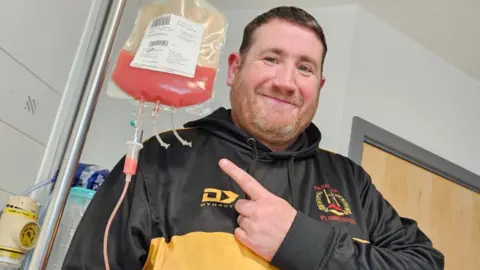 NHS Blood and Transplant A smiling man wearing a black and yellow top points at an infusion hanging from a drip stand.