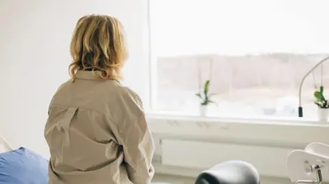 A stock image of a blonde woman sitting on a bed in a hospital room facing towards a window. She has wavy blonde hair and a heavy khaki shirt. There is a pillow covered with a blue pillowcase visible on the bed and to the right of the frame a black stool can be seen.