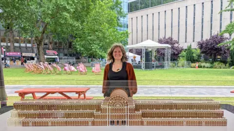 Emily Garland A woman is standing behind a gingerbread replica of Crystal Palace. She appears to be in a park, which has deckchairs and a small gazebo-type bandstand in it. There are multi-storey office buildings beyond the park.