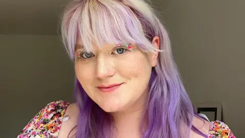 Elspeth Oakley is smiling at the camera. She has long white and purple hair and colourful eye make-up.