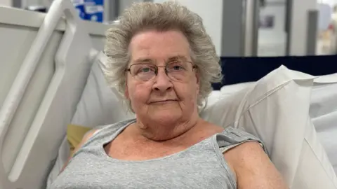 Alison Moss sits on a hospital trolley. She has grey hair and is wearing a grey vest. 