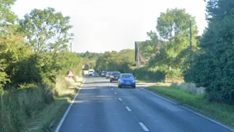 Google A Google Street View screenshot of a two-lane stretch of the A140 road in the UK, showing oncoming traffic led by a blue hatchback, with lush green hedges, mature trees, and a triangular road sign lining the countryside roadside under a bright sky.