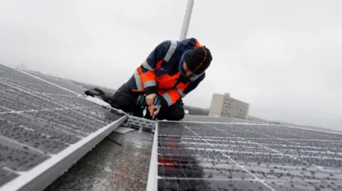 Reuters A man wearing a high-vis jacket, fitting solar panels on a roof in the rain.