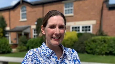Victoria Kazi, who is wearing a blue and white blouse, has dark brown hair that is tied up behind her. She is standing in front of a row of two-storey red-brick buildings that are behind bushes.