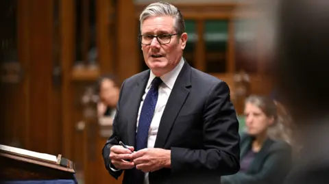 Reuters Sir Keir Starmer stands and speaks. He wears a black suit, white shirt and blue tie.