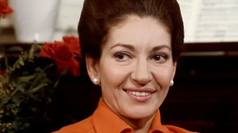 A photo of Maria Callas with her brown hair up, wearing pearl stud earrings and an orange shirt