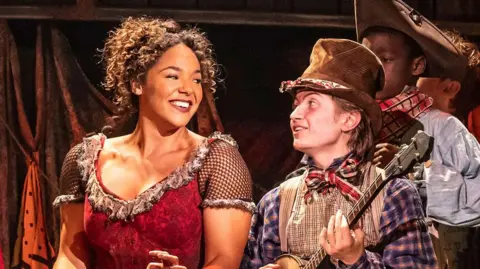 Shanay Holmes is smiling in a red Victorian dress and dancing next to an actor playing Artful Dodger 