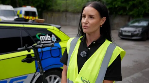 Hayley Sutcliffe from the Motor Insurers' Bureau is standing in front of a police car and an e-scooter, she has dark shoulder length hair and is wearing a high-vis vest.
