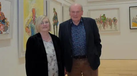 Liam McArdle A man and woman are standing in an art gallery. They are both wearing glasses and smiling at the camera. 
