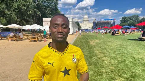 Christian Boateng stands posing in a football jersey. Behind him you can see the US Capitol and FIFA World Cup celebrations