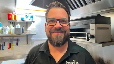 A man with short dark hair and a dark beard is standing in a commercial kitchen. He is wearing glasses and a black open-necked shirt.