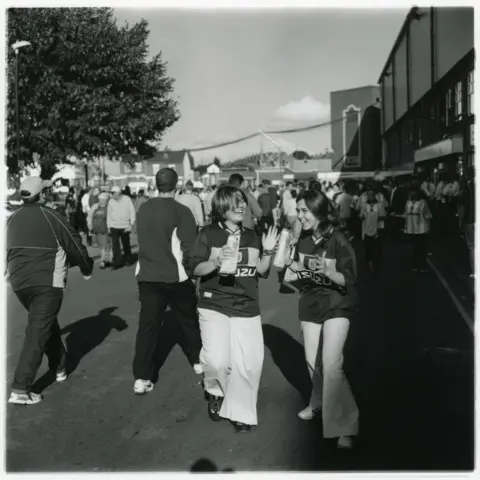 Jason Scott Tilley/Lower Block A black and white image of two female Coventry City fans outside Highfield Stadium. They are laughing. Both are wearing Coventry City tops and white trousers. 
