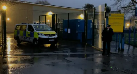 A police van is parked inside the gates of Kingsbury High School in Brent at dusk, while an officer stands nearby on a wet road with school buildings and signage visible.