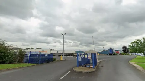 The main entrance of Wetherby-based Moores Furniture Group. It shows buildings in the distance and in the foreground blue gates and a security hut with a number of large trucks bearing the Moores logo parked behind the main gate