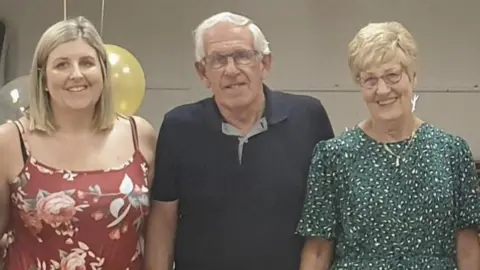 St Elizabeth Hospice A woman on the left standing next to an older man in the middle and an older woman on the right. They are all smiling at the camera. Behind them are two balloons.
