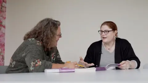 Pictured is Paula with some books. She is wearing a black cardigan and grey top. She has glasses on and silver hooped earrings. Next to her is a lady with brown hair. She has a grey top of which has colourful patches.