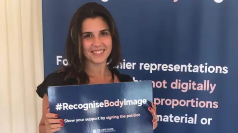 Elle Seline Elle Seline smiling, holding a sign that says #RecogniseBodyImage, show your support by signing the petition. She is standing in front of a banner that has wording on.