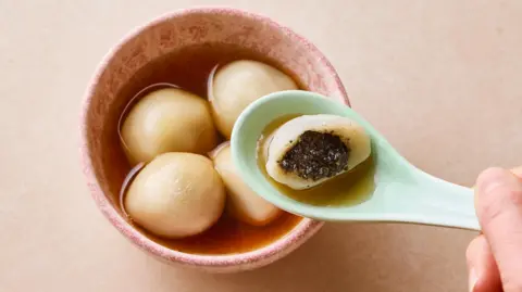 A bowl of black sesame-filled dumplings in a syrup. A hand holds a green spoon, containing a half eaten dumpling, just above the bowl
