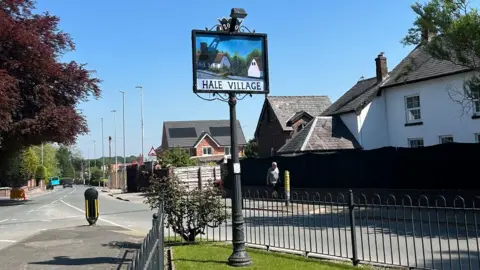 A village street scene with a road winding into the distance. A small villages green has a street sign which reads Hale Village and features a painting of the village. A white building is in the background and more modern houses can be seen in the background.