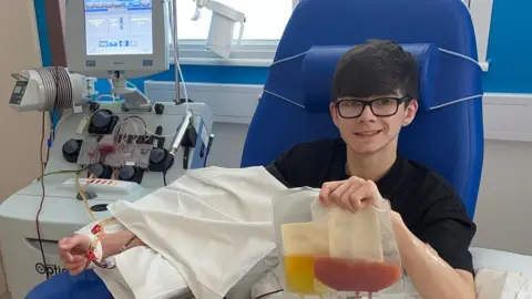 Jamie McGuigan has short dark brown hair and glasses. He is sitting on a hospital blue chair, smiling, with a machine attached to his arm. He is holding up two plastic medical bags, one filled with a red fluid, the other yellow.
