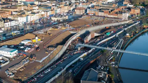 Stockton Borough Council An aerial view of a narrow, modern white footbridge over the River Tees. The bend of the river can be seen to the right with the bridge disappearing out of shot. On the left, the river gives way to yellow building machinery on the bank. In the foreground is a building with a tiled roof behind which a line of cars are parked. A road along the bank has tall silver fencing on one side and short red and white barriers on the other. There is a large expanse of flattened earth between the road and the buildings of Stockton.