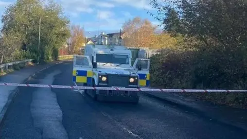 A PSNI landrover on an empty road, which has trees on either side. Its two side doors are open and it sits behind some police tape.